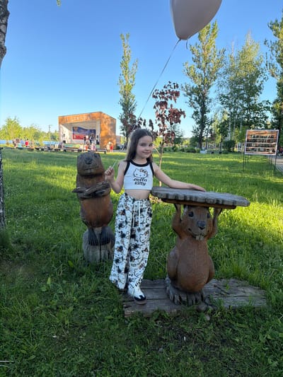 The image depicts an outdoor scene in a park in Zaprudnya, Russia, likely during a public event on Russia Day.

In the foreground, a young female child, approximately 7-10 years old, with long dark hair, stands smiling at the camera. She wears a white crop top featuring a "Hello Kitty" logo and character motifs, and white loose-fitting pants patterned with "Hello Kitty" characters and black splatters. Her footwear consists of white sneakers with black toes and soles. Her left hand (viewer's right) holds the string of a large, partially visible white balloon at the top of the frame. Her right arm (viewer's left) rests on a small, rectangular wooden tabletop supported by a carved wooden beaver sculpture to her right.

To the child's immediate left stands another carved wooden beaver sculpture, facing slightly towards the viewer, with its arms crossed over its chest, holding a stick. Both beaver sculptures are dark brown, detailed, and stand on wooden bases amidst green grass.

In the midground, a well-maintained grassy park area is visible with scattered deciduous trees. To the right, a rectangular sign on a stand displays numerous small framed photographs, identified by the text "БЕССМЕРТНЫЙ ПОЛК" (Immortal Regiment), indicating a commemorative display. A paved pathway runs horizontally behind the main subjects.

In the background, a temporary outdoor stage structure is present. The stage has a reddish-brown or orange roof and side panels. On the stage, several individuals are visible, with musical equipment such as speakers and stands. A large banner on the stage features the colors of the Russian flag (white, blue, red) and the text "С ДНЁМ РОССИИ" (Happy Russia Day), indicating a celebration for Russia Day. Beyond the stage and pathway, a body of water is visible on the left. The sky is clear and bright blue, suggesting a sunny day.