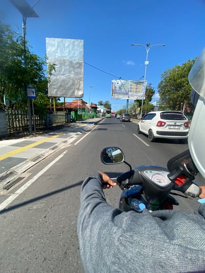 The image, taken from the perspective of someone riding a motorcycle on a sunny day in Jawa, Indonesia, shows a road with light traffic. The road has two lanes in the direction of travel, with a white line dividing them. A white Suzuki Ignis car is visible in the right lane. On the left side of the road, there is a sidewalk with tactile paving, a green fence, and a large, empty billboard structure. Further down the road, there are more billboards and trees. The sky is clear and blue, indicating good weather. The motorcycle's handlebars, dashboard, and the rider's left arm in a gray sweater are in the foreground. A water bottle is attached to the motorcycle's body.