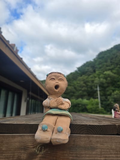 A close-up, low-angle shot shows a small, tan clay figurine of a child sitting on a wooden surface, outdoors. The figurine is depicting a child with an open mouth, as if singing or shouting, with hands clasped in front of its chest. It is wearing a light blue skirt and what appear to be two light blue decorations on its feet. The figurine has dark hair tied up with blue beads.

Behind the figurine, a dark brown wooden deck or table is visible. Further in the background, a building with a brown roofline and large windows is seen to the left. To the right and behind the deck, a densely forested green hill rises under a cloudy sky. A distant, out-of-focus figure of another figurine can be seen on the right side of the deck. The scene is likely located in Gumi-si, South Korea, given the context.