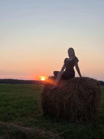 A young woman in a dark dress with a floral pattern sits on a large, round bale of hay in a field at sunset. The sun is low on the horizon, casting a warm orange glow across the sky and illuminating the field with golden light. The woman is silhouetted against the sunset, her face partially obscured by shadow. She is barefoot, with her legs bent and crossed at the ankles. The hay bale is rough and textured, composed of dry straw. In the background, a line of trees forms a dark silhouette against the colorful sky. Other hay bales are visible in the distance, scattered across the green field. The overall scene depicts a serene and picturesque rural landscape during twilight.