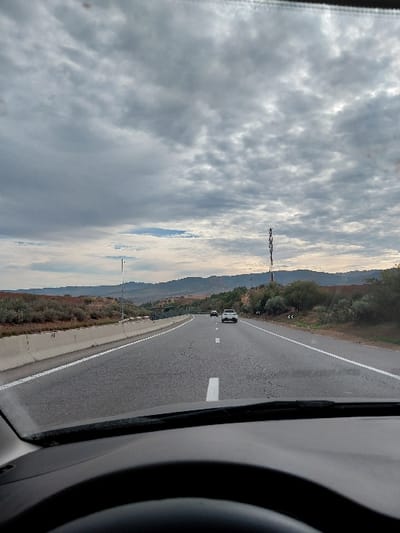 A highway view is captured from within a car, likely the driver's perspective. The road stretches ahead, marked with white lines and sparse traffic. Rolling hills line the horizon under a cloudy sky. A tall radio tower stands prominently against the backdrop. Part of the car's interior, including the dashboard and steering wheel, frame the scene, hinting at a journey underway.
