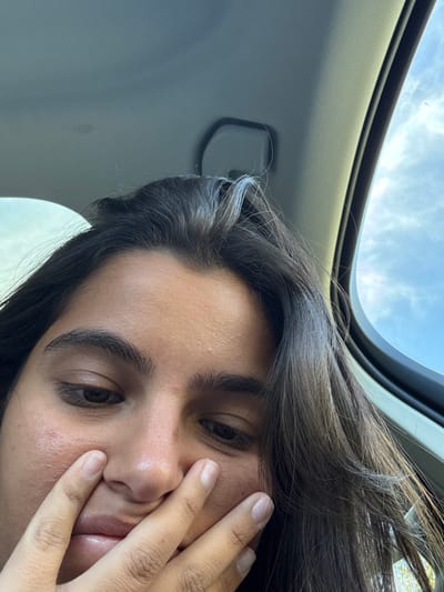 The image displays an individual seated inside a vehicle, located in the city of Princeton, United States. The subject, seen from an upward angle in a close-up, has dark, slightly wavy hair that partially obscures the forehead and sides of the face. Their dark eyes are cast downwards, and the left hand covers the lower portion of the face, obscuring the mouth and chin. Visible environmental elements include the vehicle's light-colored headliner in the upper left, a dark overhead grab handle, and a sunroof or window frame on the right side, revealing a blue sky with scattered white clouds, indicative of daytime.