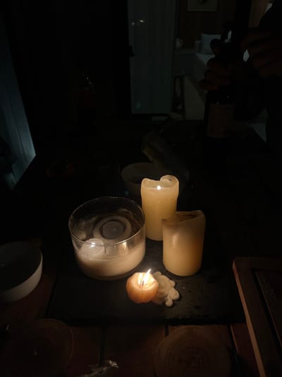 The scene is dimly lit with candlelight illuminating a tabletop arrangement. Several partially burnt candles, including one encased in a glass holder, are grouped together. In the background, hands are seen holding a wine bottle and corkscrew. Sliced wooden discs are arranged vertically against a dark wooden surface on the right side. A white bowl sits at the edge of the table. The overall atmosphere is dark and intimate.