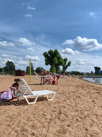 The image depicts a sunny day at a beach, likely in the city of Myadzyel, Belarus, given the context of the location tag. In the foreground, two white plastic lounge chairs are visible on the sand. One is occupied by a person wearing a brown top and blue shorts, who appears to be sitting on a blanket and has a pink bag nearby. The other lounge chair is empty. Further back, a group of people are gathered around a table. A man with a bare chest and shorts is standing, while others are seated. The beach is populated with other individuals, some are walking, others are sunbathing, and some are in the water. The water appears to be a lake or a designated swimming area, with people swimming and some in inflatable rings. The environment includes sand, trees, and a clear blue sky with scattered white clouds. In the background, there are buildings and a white gazebo structure.