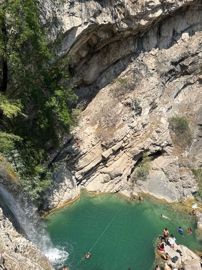 The image shows a steep, rocky cliffside near a body of water. The cliff is mostly gray and brown, with some patches of green vegetation. At the top right of the image, people are swimming and gathering near the water's edge. A waterfall cascades down from the top right, creating a splash at the bottom. The water is a clear, turquoise color. A line stretches across the water, and people are swimming in the water. The location is San Miguel Atlapulco, Mexico.