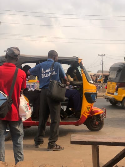 The image depicts a street scene in AlakIa, Nigeria, under an overcast sky. In the foreground, three male subjects are present. On the far left, a man is seen from behind, wearing a red short-sleeved shirt, a grey baseball cap, light blue jeans, and carrying a black cross-body bag. He holds a white plastic bag with red and black graphics in his left hand. To his right, a second man, also viewed from behind, wears a blue short-sleeved shirt with "ALWAYS ORIGINAL" printed in white across the back. He wears dark trousers and brown sandals, and a black messenger bag hangs from his left shoulder.
This man stands next to a yellow and red three-wheeled commercial passenger vehicle, a 'keke napep'. The vehicle features a yellow body with red accents and stripes on the lower front, including a circular yellow sticker with a black '90'. The driver, a male wearing a yellow shirt and dark trousers, is seated inside the keke napep, visible from the chest up and facing forward. A dark canopy covers the vehicle's passenger section.
In the mid-ground to the right, another yellow keke napep with a black canopy is visible. The background features utility poles supporting multiple overhead power lines against the light grey sky, with indistinct figures and structures further down what appears to be a dusty or unpaved road. A portion of a dark wooden bench or table is visible in the lower right foreground.
