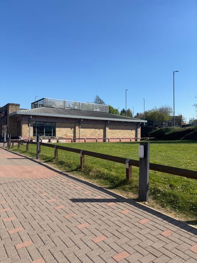 The image shows a paved walkway leading to a wooden fence and a grassy area. The walkway is made of interlocking grey bricks with reddish-brown accents. The fence is made of wooden posts and rails. Behind the fence is a grassy area with some small yellow flowers. The sky is a clear blue. On the left side of the image, a single-story brick building with a flat roof and a metal grate is visible. There are no people present. The image appears to have been taken on a sunny day. The location is Bradford, United Kingdom, specifically the area around the coordinates latitude: 53.791, longitude: -1.748. The building might be a retail outlet or restaurant.