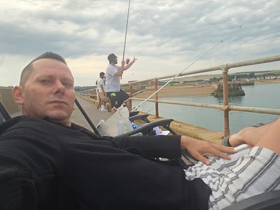 A man is taking a selfie while sitting in a black reclining chair on a pier. He is wearing a black textured sweater and has short, buzzed hair. To his right, another person is standing with a fishing rod. In the background, several other individuals are seen fishing. The pier is made of concrete and has a rusty metal railing. The sea is visible to the right, and beyond that, a beach and buildings line the shore. The sky is cloudy. The location is identified as Adur, United Kingdom.