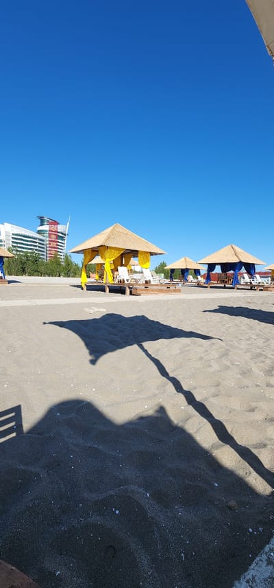 The image shows a bright, sunny day at a beach in Türkmenbaşy, Turkmenistan. In the foreground, dark sand with small white shells is visible. The shadow of a large beach umbrella, complete with its pole, stretches across the sand from the left side of the image. Further back, several thatched-roof cabanas are set up on wooden platforms. The cabana on the left is adorned with yellow fabric curtains and has white lounge chairs underneath. To its right, another cabana is visible with blue fabric curtains. In the background, a large, modern building with a curved glass facade and a red and gold patterned section stands amidst green trees. The sky is a clear, vibrant blue, indicating fair weather. The scene depicts a tranquil resort or public beach area.