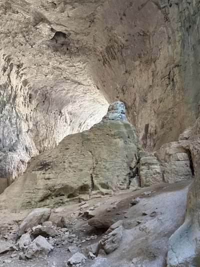 The image displays the interior of a cave, likely in Karlukovo, Bulgaria, characterized by rough, textured rock formations and a high, arched ceiling. The lighting suggests daylight entering from an opening, illuminating the cavern and casting shadows. Dominating the center is a large, irregularly shaped rock pillar, which appears to be a significant geological feature within the cave. The ground is covered in loose rocks and dirt, indicating a natural, undeveloped environment. The cave walls exhibit layering and erosion, suggesting a long geological history. The overall impression is one of natural grandeur and geological timescale.