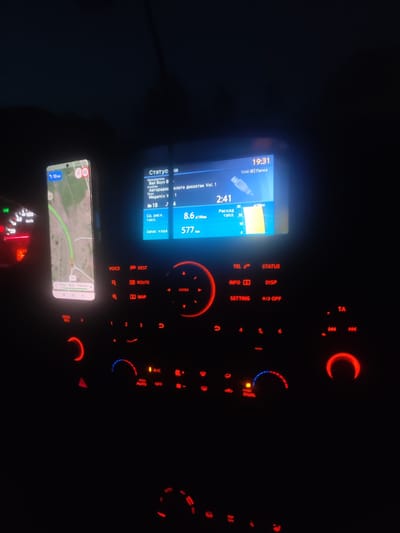 This media file captures a nighttime scene inside a car. The focus is on the car's dashboard, which is illuminated by the glow of various screens and buttons. On the right side of the dashboard, a car's infotainment system displays song information, including the artist "Bad Boys B" and the song title "Megamix V.1". It also shows the current time as 19:31 and a fuel consumption reading of 8.6 L/100km, with a remaining range of 577 km.
To the left of the infotainment screen, a smartphone is mounted, displaying a navigation app with a route highlighted in green, indicating a journey in progress. The phone shows the distance to the destination as 10 km. Also visible on the dashboard is the car's speedometer, showing a speed of approximately 60 km/h, and the fuel gauge.
The surrounding environment is dark, suggesting it is either evening or night. The overall atmosphere is functional, with the car's controls and displays being the primary subjects. The red and blue backlighting of the dashboard buttons adds to the nighttime ambiance. There are no people visible in the frame. The visible text is primarily in Russian, pertaining to car functions and navigation.