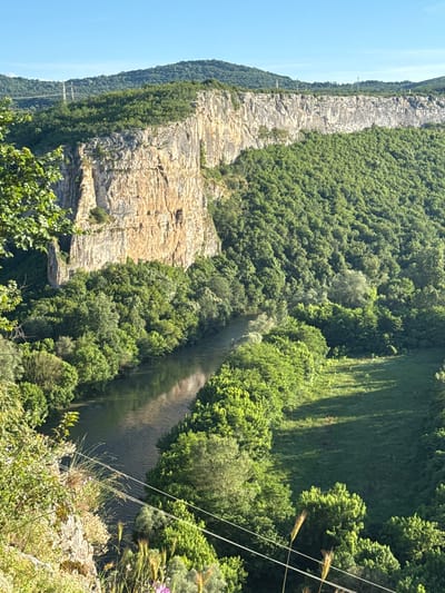This is a high-angle, full outdoor shot of the Karlukovo Karst region in Bulgaria, featuring the Iskar River. A sheer, light-colored rock cliff face dominates the left side of the image, with its textured surface catching the sunlight. Dense, vibrant green foliage covers the slopes of the hills and extends along the riverbanks, creating a lush, natural landscape. The Iskar River flows gently through the scene, its surface reflecting the sky and surrounding trees. In the distance, rolling hills covered in trees form the horizon under a clear blue sky. Power lines are visible traversing the landscape in the upper left and lower right portions of the frame. There are no people or animals depicted in the image.