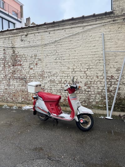 The scene depicts an outdoor urban environment, specifically a street or alleyway in Ixelles - Elsene, Belgium, under an overcast sky with recent or ongoing light snowfall. A red and white/silver Honda Lead scooter is parked facing right on a dark, wet asphalt surface, supported by its kickstand. The scooter's seat is red, and a white storage box is affixed to the rear rack. Light snow accumulation is visible on the scooter's handlebars, front fender, footrest area, and seat. A distressed, light-colored brick wall with significant peeling paint, revealing red bricks, forms the primary background. Horizontal metal conduits run along the upper portion of the wall, which also has a light dusting of snow on its coping. To the left, above the wall, parts of a modern building with a white facade and dark windows, and a residential building with a pinkish-orange facade and a black balcony railing are visible. To the right, two vertical silver-colored tubular poles with circular bases stand near the wall, also bearing traces of snow. Patches of snow and sparse icy vegetation are present along the base of the wall and on the asphalt. No individuals are present in the image.