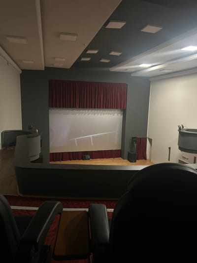The image depicts an indoor auditorium or cinema space. The foreground shows the backs of two dark-colored seats, one partially obscured by the other. Beyond the seats, a dark-colored stage area is visible with a large projection screen at the back. Red curtains frame the top and bottom of the screen. To the left of the stage, a curved, dark gray podium or control desk is present, with a small item, possibly a bottle, on its surface. To the right of the stage, a black speaker unit is positioned near red curtains. The ceiling features a dual-tone design, with dark gray sections and white sections containing recessed square lighting panels. White blinds are visible on the left wall. The scene suggests a formal presentation or performance venue, likely located in Byala Slatina, Bulgaria, given the prompt's context. There are no people clearly visible in the image.