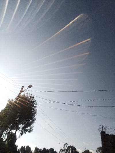 The image presents an outdoor scene in Nachu ward, Kenya, under a clear, light blue sky. A strong sun glare emanates from the lower-left, manifesting as multiple parallel, diagonal streaks of bright light across the sky. In the lower-left quadrant, a dark utility pole supports a singular street light fixture, partially obscured by the dense green canopy of several trees. Multiple dark electrical or communication lines traverse horizontally across the mid-ground and upper-mid-ground. In the lower-right corner, a dark, solid structure, consistent with a wall or fence post, is topped with coiled barbed wire. No individuals, specific actions, or interactions are observable.