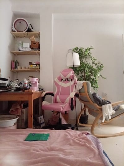 The image depicts an indoor scene, possibly a bedroom or living area, characterized by a collection of objects suggesting a lived-in space. A prominent pink and white gaming chair is positioned in the center-right of the frame. To its left, a wooden desk is cluttered with various items, including what appears to be a small pink sewing machine, bottles, and other miscellaneous objects. Above the desk, a shelf unit displays more items, such as books, containers, and a basket.
In the background, to the right of the gaming chair, a rocking chair with clothing draped over it is visible, and behind it, a large artificial plant stands beside a floor lamp with a white shade. In the foreground, a light pink blanket or throw covers a surface, with a green notebook resting on it. The walls are painted white, and the flooring appears to be light-colored tile or linoleum.
This scene does not provide any specific visual cues that would allow for its localization in Olula del Río, Spain, or any other city. The interior setting is generic, lacking architectural or decorative elements that are distinctly Spanish or specific to that particular municipality.