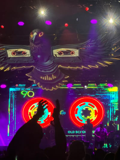 The image depicts a concert stage with a large screen displaying a stylized bird with cassette tapes for wings, and the text "90" and "OLD SCHOOL". The bird graphic is purple and yellow. The screen features neon lights and a digital pattern. In the foreground, silhouettes of concert-goers are visible, with hands raised. Several musicians are on stage, one playing a guitar. Purple and white stage lights illuminate the scene. The image appears to be taken at the Veterans United Home Loans Amphitheater in Virginia Beach, United States.