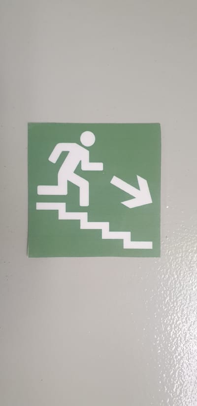 An emergency exit sign is affixed to a light gray wall. The sign is square with a green background and a white pictogram. The pictogram depicts a stylized person running down a set of stairs, indicated by three descending horizontal bars. To the right of the stairs, a white arrow points diagonally downwards and to the right, indicating the direction of the exit. The sign is located on a wall in Rooppur, Bangladesh, likely within a building associated with the Rooppur Nuclear Power Plant. The wall appears to be painted or finished with a slightly textured surface.