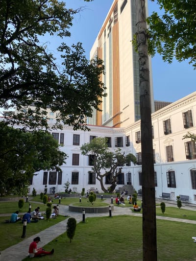 The image depicts a courtyard with a white, multi-story building and a tall, modern building in the background. The courtyard is covered in lush green grass with paved pathways. Several people are scattered throughout the courtyard, sitting on the grass and engaged in various activities such as reading or studying. Some are alone, while others are in small groups. The building in the foreground features numerous open windows with dark wooden frames. The overall scene suggests a peaceful and studious atmosphere, likely within an educational institution or a similar public space in Kathmandu Metropolitan City, Nepal, given the architectural style and landscaping. The lighting indicates it is daytime, with clear sky visible.