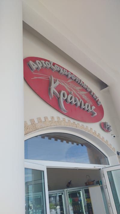 The image shows the exterior of a bakery or pastry shop in Nea Michaniona, Greece. A large, red, oval sign with silver, raised lettering is mounted on the wall. The Greek text on the sign appears to be "Αρτοζαχαροπλαστείο Κρανάς" which translates to "Bakery Pastry Shop Kranas". A stylized wheat stalk is depicted behind the word "Κρανάς". Below the sign, a decorative border of light-colored stone with a crenellated pattern runs along the top of an arched window. The window is made of glass and reflects the sky. French doors, also made of glass, are partially open, revealing refrigerators inside the establishment, stocked with various drinks and products. A small security camera is visible on the wall to the right of the arched window. The overall scene depicts the entrance to a commercial establishment in a Greek coastal town.