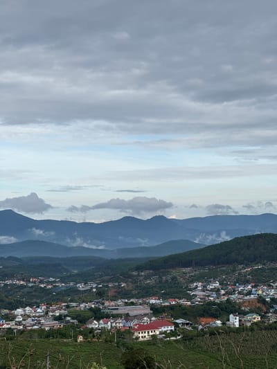 An elevated perspective showcases Trường Thọ, Vietnam. Multiple dark mountains line the center of the frame. A cloudy sky occupies the left side. The city is on the right, featuring clustered buildings interspersed with trees.