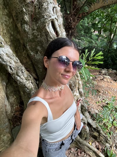 In a selfie taken outdoors in a lush, green environment, a young Caucasian woman with dark hair and sunglasses poses next to a large tree trunk. She is wearing a white, off-the-shoulder top and grey denim shorts. A floral choker adorns her neck. The background reveals a dense jungle with foliage and rocky terrain, consistent with a location in Thuận Nam, Vietnam.
