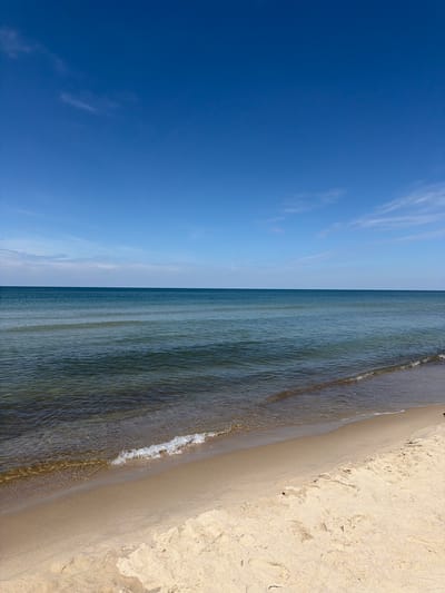 The image shows a serene beach scene on a clear day. The foreground is dominated by light-colored sand, with some subtle footprints visible. A gentle wave, with white foam, is washing ashore, creating a clear line between the wet and dry sand. Beyond the sand, the calm, shallow water transitions to a deeper, bluish-green hue. The horizon line is sharp and distinct, separating the sea from a clear blue sky. A few wispy clouds are scattered across the upper portion of the sky. The scene depicts the natural beauty of a coastal environment, likely the Baltic Sea coast near Morskoye, Russia, given its tranquil and expansive appearance. There are no people or man-made objects clearly visible in the frame, contributing to the peaceful atmosphere.