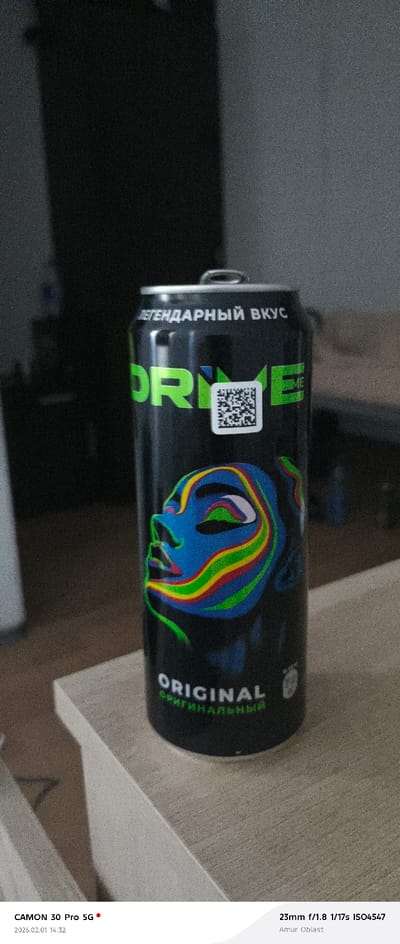 A slightly grainy, close-up shot focuses on a black can of Drive Me energy drink. The can's design features a colorful, abstract face with blue skin and rainbow-like stripes, along with a prominent QR code. Text in Cyrillic, "Легендарный Вкус" (Legendary Taste) and "Original", adorns the can. The can sits on a light-colored wooden surface. The background is dark and indistinct. Camera information along the bottom indicates the image was taken with a CAMON 30 Pro 5G at 23mm f/1.8, 1/17s, ISO4547, in the Amur Oblast region.