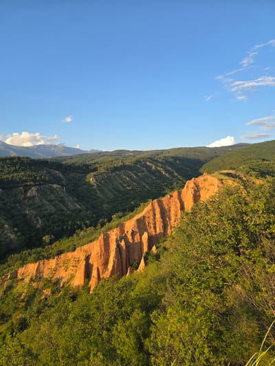 The image displays a panoramic natural landscape in Stob, Bulgaria, characterized by prominent geological formations under a clear blue sky. The central feature is a series of reddish-orange earth pyramids, known as the Stob Pyramids, extending diagonally from the mid-left to the mid-right. These eroded formations exhibit vertical striations and spire-like structures, intensely illuminated by low-angle sunlight, casting warm tones. Dense green deciduous and coniferous vegetation covers the foreground, with bushes occupying the lower right corner, and extensively throughout the midground valleys and slopes surrounding the pyramids. In the background, forested mountains with dark green trees stretch across the horizon, culminating in distant, snow-capped peaks on the far left. The sky is bright blue, featuring scattered white cumulus and cirrus clouds. No human subjects, animals, or artificial structures are visible, indicating an untouched natural environment.