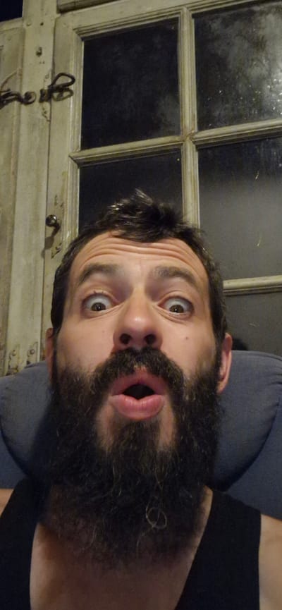 A man with a dark beard and dark hair, who appears to be in his late 30s or early 40s, is captured in a close-up selfie. His eyes are wide open, and his mouth is pursed open, giving him a surprised or shocked expression. He is wearing a black tank top. The man is seated, leaning against a dark blue padded chair or headrest. Behind him, a rustic wooden door with a metal latch is visible, and beyond that, a multi-paned window with dark, opaque glass. The scene appears to be indoors. The location is stated to be Wattenwil, Switzerland.