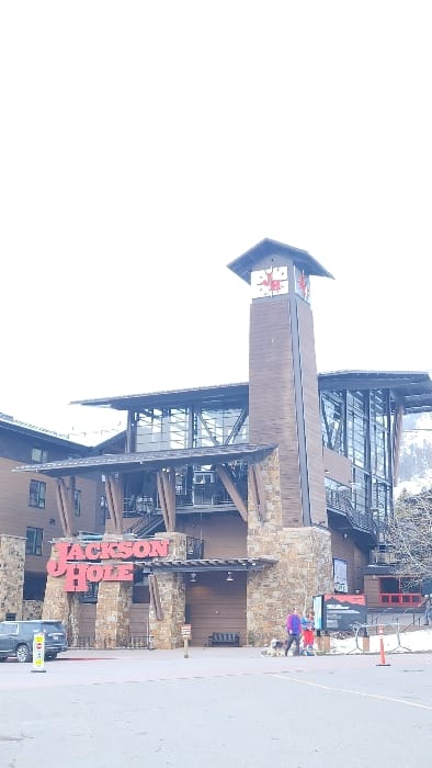 An exterior, eye-level, wide-angle photograph depicts the base lodge of Jackson Hole Mountain Resort in Teton Village, Wyoming, United States of America, under a bright, overcast sky. The multi-story building features a rustic-modern architectural style, characterized by stone masonry bases, dark wood paneling, and large glass window sections. A prominent red illuminated sign spelling "JACKSON HOLE" is affixed above a covered entrance on the left side of the structure. To the right, a tall, slender clock tower constructed of wood and stone rises, topped with a sloped roof. Four clock faces display red numerals and hands against a white background, centered with a red "JH" logo. The visible clock face indicates approximately 9:15.
In the mid-ground, a paved surface, likely a parking area or plaza, occupies the foreground. Patches of snow are present on the ground adjacent to the building, particularly on the right side. A dark grey SUV is parked on the left of the paved area, its rear facing the viewer, positioned near a yellow "STOP" sign on a pole.
On the right side of the paved area, in front of the building, a group of three individuals and one large shaggy-haired dog are standing on a mix of pavement and snow. One person wears a purple jacket, another a red jacket, and a third is partially obscured. They appear to be stationary. An orange traffic cone is positioned further right on the paved surface. Information boards are visible behind the individuals, against the building facade. Distant, snow-covered mountains are faintly visible in the background behind the building on the right.