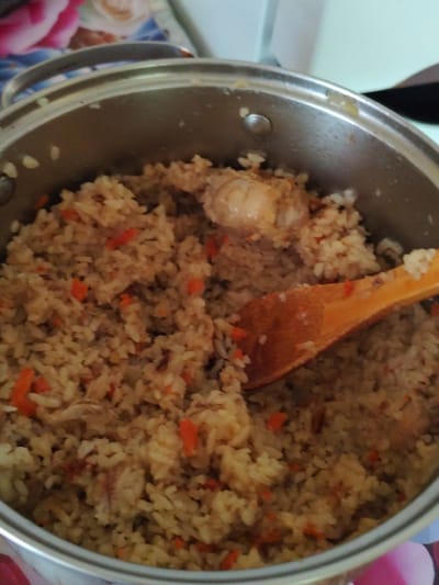 A close-up, overhead shot shows a metal pot filled with pilaf, a rice dish with meat and vegetables. A wooden spoon rests on top of the pilaf, its handle extending out of the pot. The pilaf appears to be cooked with rice, diced carrots, and pieces of meat. The lighting is natural, coming from above, and casts soft shadows on the food. The background is blurred, but hints of floral patterns suggest a domestic setting. The image quality is moderate, with some graininess and slight focus issues. The scene is located in Shakhtarsk, Ukraine.