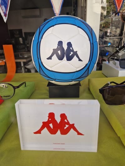 The image displays a store window display featuring a Kappa branded soccer ball and other merchandise. The soccer ball is white with blue pentagons and the Kappa logo, two silhouettes of people sitting. In the foreground, a white box with the red Kappa logo is visible. To the left, a pair of glasses with the Kappa logo on the lens is displayed on a green surface. To the right, a pair of sunglasses is also visible. In the background, shelves with various items, possibly eyewear, and a window showing an outdoor environment can be seen. The store is located in Antony, France, as indicated by the presence of theKappa brand, a popular sportswear company.