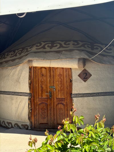 The image shows the door of a yurt, a traditional dwelling used by nomadic people in Central Asia, in Tong, Kyrgyzstan. The door is wooden, with carved decorative designs, and is secured with a metal latch and key. Above the door, on the yurt's outer covering, is a brown diamond-shaped sign with the number "2" in black. The yurt's covering is a thick, felt-like material with decorative patterns along the edges. To the right of the door, a plant with green leaves and red-tinged buds is visible, partially obscuring the entrance. A section of a white structure, possibly a building or vehicle, is visible on the left side of the frame.