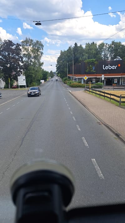 The image shows a view from a vehicle traveling down a road in Kreuztal, Germany. The road is asphalt with white dashed lines indicating lanes. On the right side of the road is a sidewalk made of red bricks, a wooden fence, and a grassy area. Beyond the fence is a building with a dark facade and the name "Leber" prominently displayed in white letters. Trees line the street, and the sky is partly cloudy. There are a few cars visible on the road. In the foreground, the top of a dashboard or camera mount is blurred.
