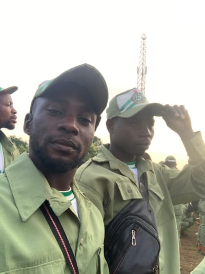 Two adult males, likely members of the National Youth Service Corps (NYSC), are prominently featured in an outdoor close-up photograph taken in Kubi A, Nigeria. The man on the left, with a dark complexion, full beard, and mustache, wears an olive-green uniform shirt over a white and green striped undershirt, and an olive-green cap. He gazes directly towards the camera. To his immediate right, another male, also with a dark complexion and dressed in an identical olive-green uniform shirt, wears a light green and white NYSC cap; he is seen adjusting the brim of his cap with his right hand. A black shoulder bag with silver zippered compartments is slung across his chest. In the background, partially obscured individuals in similar olive-green uniforms are visible, indicating a larger gathering. A tall, metallic lattice-structure cellular communication tower rises distinctly against the bright, hazy sky. The environment consists of light-colored ground and distant, indistinct green foliage along the horizon.