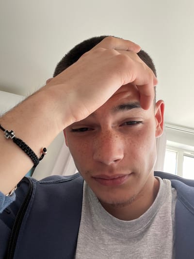 A young man with a short haircut, fair skin, and freckles is looking at the camera from a tilted angle. He is wearing a grey shirt and a dark blue hooded jacket. His left hand is raised to his forehead. He is wearing a black braided bracelet with a silver cross and bead. The background is a white wall with a window. The image location is not specified within the provided image.