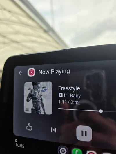 The image shows a close-up of a car's infotainment screen displaying music playback information. The song playing is "Freestyle" by Lil Baby, and the time elapsed is 1:11 out of a total song length of 2:42. The album art is visible, along with playback controls like pause, skip, and like. The display also shows the time as 10:05. The location is impossible to ascertain. There are no details indicating the city of Houghton Regis in the United Kingdom.