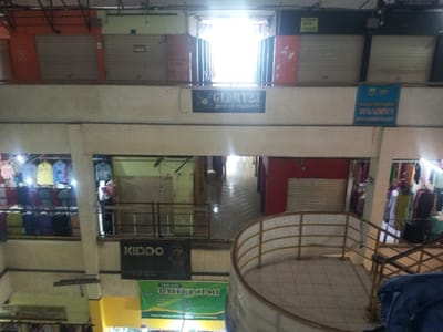 A multi-level indoor market shows closed shops with roller shutters, suggesting inactivity. Clothing racks displaying colorful garments flank a central walkway bathed in strong sunlight. Signage includes "GLORY23", "KIDDO", and a banner with "BALE BUMI". The architecture features a tiled floor, curved railing, and a mix of open and closed shopfronts, creating a layered visual perspective within the building.