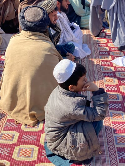 Several people lie on a woven mat outside in the sun. The individuals appear to be Middle Eastern, with some wearing traditional head coverings. A boy wearing a white skullcap and a gray jacket reclines near the foreground with his hand near his mouth. He lies next to a man covered by a tan blanket. Other men in turbans and white clothing are scattered around them. The patterned mat provides a colorful background to the group.