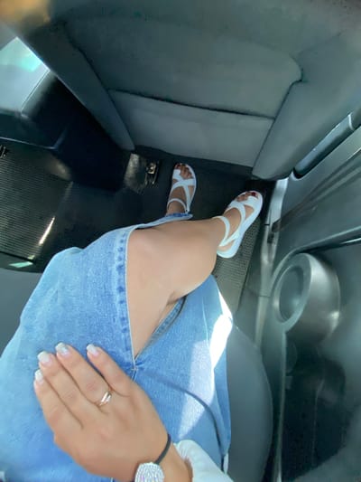 The image shows a person's legs and hands inside a car. The person is wearing white sandals and a denim skirt. Their hand is resting on their leg, and they are wearing a ring on their finger. The car's interior is visible, including the seats and a speaker. The location is the city of Tyulyugul, Russia.