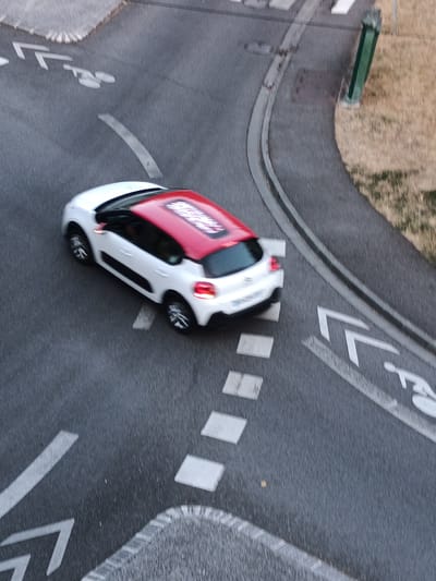 The image shows an aerial view of a white car with a red roof driving on a road in Évreux, France. The road has various markings including a bike lane symbol, pedestrian crossing symbols, and directional arrows. The car is in motion, indicated by the motion blur. The surrounding environment appears to be an urban area with a grassy verge and a green lamppost visible.