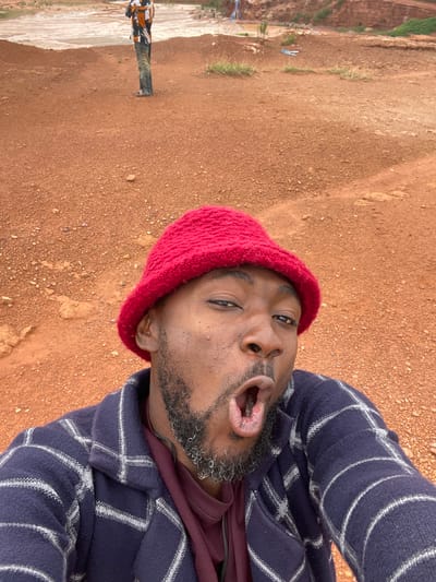 A man in a red knit hat and a plaid jacket is taking a selfie in an outdoor, dusty, reddish-brown environment, possibly a mine or construction site, with a body of water in the background. His mouth is open in an exaggerated expression. In the background, another person is standing, appearing to be looking out at the water. The ground is uneven and covered in small stones and dirt.
