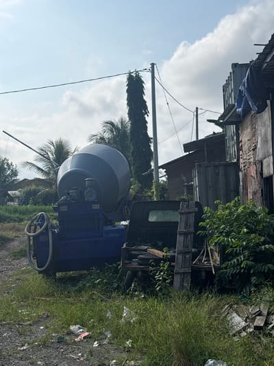 The image depicts an outdoor scene in Tampaksiring, Indonesia, characterized by a blue concrete mixer in the foreground. The mixer is situated on a gravelly path, surrounded by overgrown grass and scattered litter. Behind the mixer, a dark-colored truck bed is visible, along with various construction-related debris and wooden structures. Further in the background, palm trees and other vegetation suggest a tropical or subtropical environment. Several buildings, appearing somewhat dilapidated and constructed from corrugated metal and brick, line the right side of the frame, with power lines and utility poles visible overhead. The sky is partly cloudy. There are no people visible in the image.