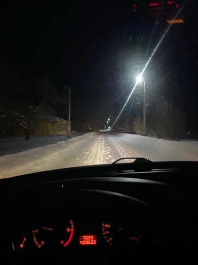 The image captures a nighttime scene from the interior of a vehicle, looking out through the windshield onto a snow-covered road. The vehicle appears to be stationary or moving very slowly, indicated by the dashboard readings.
The scene outside the car is dominated by darkness, broken by intense artificial light sources. Several bright streetlights are visible on the left side of the road, casting strong, diagonal beams of light across the snowy surface. The road itself is covered in a thick layer of snow or ice, showing distinct patterns, possibly tire tracks. In the far distance, barely discernible in the shadows and snow, are some faint structures, possibly buildings, illuminated only minimally by the distant lights.
On the right side of the frame, a portion of the car's dashboard and instrument cluster is visible, illuminated with a warm orange-red glow. A digital display is prominent, showing the numbers "409533" (likely an odometer reading) and "70.33" (possibly a trip meter reading). Above these numbers, the text "EESGOLL" is displayed. Below the digital readings, an analog fuel gauge indicates that the fuel level is nearing empty, with the needle close to the "E" mark. Other gauges, including what appears to be a speedometer with a needle near zero, are also present but less distinct. A section of the windshield wiper mechanism is visible on the far right edge of the frame.
The overall impression is of driving or being stopped in challenging winter conditions during the night.
