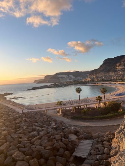 The image shows an elevated view of Amadores Beach in the municipality of Mogán, Gran Canaria, Spain. To the left is a mountain. In the center is the crescent-shaped beach with a sandy area, water, and rows of sun loungers and parasols. Palm trees line the promenade along the beach. A stone wall and walkway are visible on the right side. The sky is light blue with scattered clouds, suggesting sunrise or sunset.