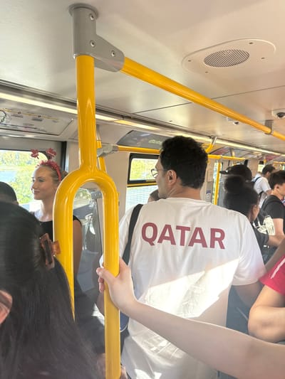 The image depicts the interior of a public transit vehicle, likely a SkyTrain in New Westminster, Canada, judging by the map displayed and the typical design of SkyTrain interiors. A group of passengers are visible, most of whom are standing and holding onto yellow handrails.
In the foreground and center, a man is seen from the back wearing a white t-shirt with the word "QATAR" printed in large, maroon lettering. He is looking to his left. To his left, a woman is visible from the chest up, facing forward and looking to her left. She has blonde hair pulled back and is wearing a red headband with decorative elements that resemble maple leaves. Her attire appears to be a black top.
Other passengers are partially visible around them, including a person on the far left with dark hair and an orange hair clip, and individuals further back wearing dark clothing and white t-shirts. The environment is consistent with a train car, featuring ceiling panels, handrails, ventilation grilles, and windows showing a blurred outdoor scene with greenery and a body of water. The overall lighting suggests it is daytime. The atmosphere appears to be one of everyday commuting.