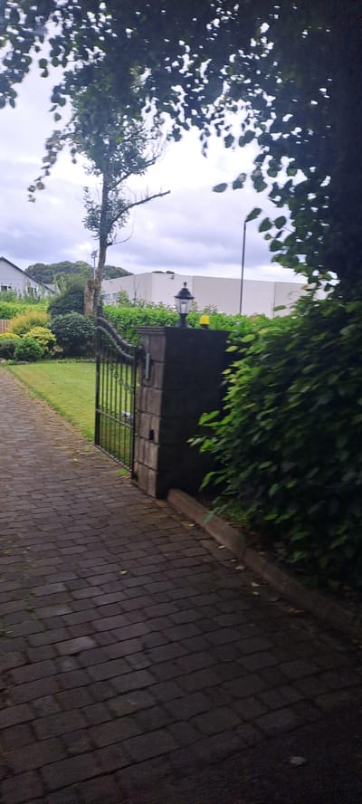 The image shows a paved driveway leading to an open black metal gate, part of a garden gate. The gate is flanked by a dark grey stone pillar on the right, topped with a black lantern with a yellow light underneath. Beyond the gate and pillar, a neatly trimmed green hedge runs along the right side of the image. To the left of the gate, a well-maintained lawn is visible, with various shrubs and trees in the background. In the far background, a white building with a flat roof and windows is partially obscured by greenery, and to the left, a residential house with a grey roof and white walls can be seen. Overcast sky with some tree branches and leaves in the foreground partially obscure the view. The scene is outdoors and appears to be a residential property.