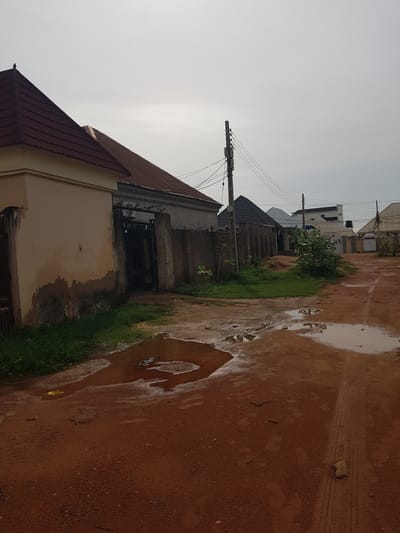 The image depicts a rural or semi-urban street scene in Wauro Jabbe, Nigeria, after a rainfall. The ground is a reddish-brown dirt road, wet and uneven, with several puddles reflecting the overcast sky. Tire tracks are visible in the mud, indicating recent vehicular activity.

Along the left side of the road, a row of buildings is visible. The foreground features the roof and upper portion of a house with a dark, textured roof and cream-colored walls. Adjacent to it are more houses, some with metal roofs and concrete or plastered walls. One building appears to have a dark, barred entrance with an air conditioning unit visible above it. Further down the street, more structures, including multi-story buildings, are present, some with visible electrical wires and utility poles.

A patch of green grass runs alongside the road between the buildings and the puddles. A small tree with dense foliage is also present. The sky is a uniform, pale gray, suggesting an overcast day, possibly morning or late afternoon, with diffused light. There is no visible text in the image. The overall impression is one of a simple, residential area, impacted by recent weather.
