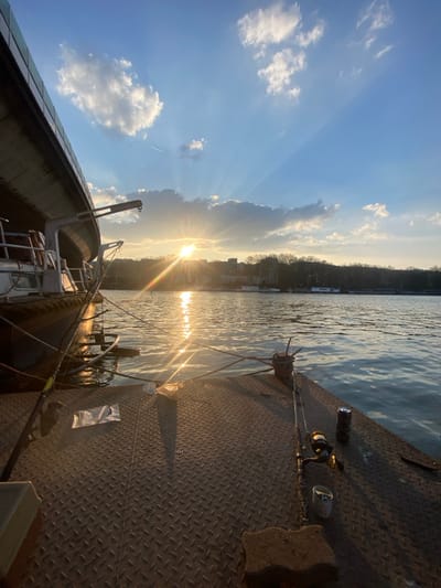 The image displays a waterside scene on the River Seine in Boulogne-Billancourt, France, during golden hour, characterized by low sun exposure. The foreground features a textured metal surface, likely a dock or deck, on which two fishing rods are laid horizontally. Adjacent to the left rod is a small, crumpled white plastic bag, while a dark beverage can and a small white cylindrical container are positioned near the right rod. A metal mooring cleat is visible centrally on this surface, and a brown rectangular block rests in the lower right corner. To the left, a section of a dark-hulled vessel or barge is docked, secured by ropes to the foreground structure, and an attached davit or crane mechanism is discernible on its side. The midground is dominated by the river's surface, reflecting a bright, direct sun glare that forms a shimmering path across the water. The background reveals the opposite riverbank, lined with deciduous trees (appearing bare), behind which urban buildings are visible. Several smaller boats or structures are present on the distant water. The sky is partially cloudy blue, with the prominent low sun emitting distinct crepuscular rays through and around scattered white cumulus clouds. A faint aircraft contrail is visible in the upper left sky quadrant. No individuals are present in the frame.